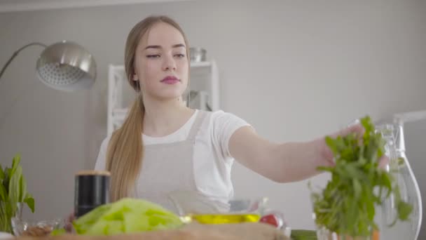 Blonde confiante fille caucasienne prenant des feuilles biologiques vertes pour la salade et les mettre sur la planche à découper. Portrait d'une jeune femme sérieuse cuisinant des aliments diététiques sains à l'intérieur. Mode de vie sain . — Video