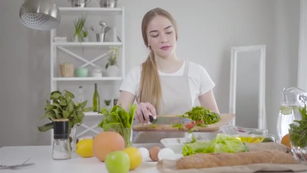 Portret skupionej młodej dziewczyny dodającej do miski pokrojone składniki. Biała kobieta przygotowuje sałatkę z pomidorów, ogórków i zieleni. Dieta, wegetarianizm, zdrowe odżywianie, styl życia. — Wideo stockowe