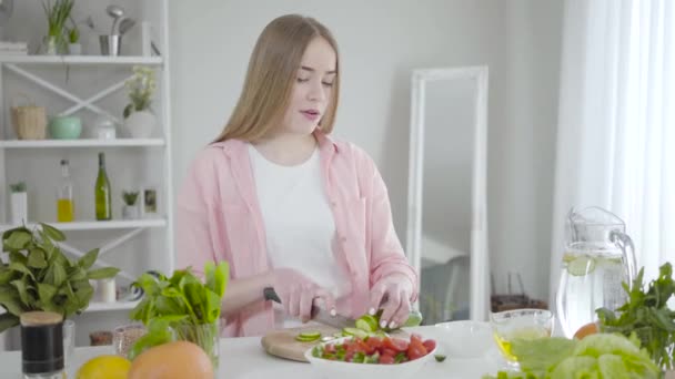 Joven mujer caucásica alegre rebanando pepino, bailando y cantando, Retrato de chica rubia alegre divirtiéndose como cocinar deliciosa ensalada orgánica en el interior. Alegría, estilo de vida saludable, felicidad . — Vídeos de Stock