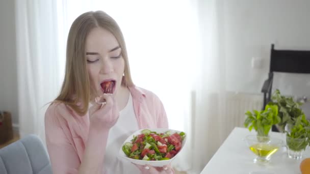 Retrato de una mujer caucásica feliz comiendo ensalada fresca de primavera de tomates, pepinos y verdor. Delgado hermosa chica disfrutando de sabrosa comida vegetariana en el interior. Estilo de vida saludable, vegetarianismo . — Vídeo de stock