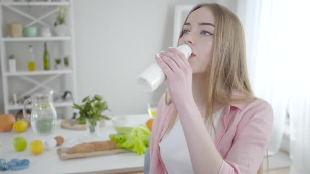 아름다운 금발의 백인 여성 이 자 헤이즐의 눈으로 유기농 요구르트를 실내에서 마시고 있습니다. 건강에 좋은 청량 음료를 즐기는 행복 한 소녀의 모습. 생활 방식, 만족, 광고. — 비디오