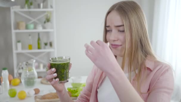 Δυσάρεστο λευκό κορίτσι κλείνει τη μύτη με τα δάχτυλα για να πιει πράσινο υγιεινό κοκτέιλ. Πορτρέτο της λεπτής νεαρή ξανθιά γυναίκα με υγιεινό ποτό σε εσωτερικούς χώρους. Τρόπος ζωής, υγειονομική περίθαλψη, δίαιτα. — Αρχείο Βίντεο