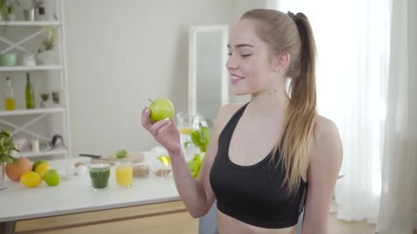 Wanita muda Kaukasia sportif menggigit apel hijau dan tersenyum di depan kamera. Potret gadis kurus ceria dalam pakaian olahraga makan buah organik di pagi hari di rumah. Joy, gaya hidup, keindahan, kebahagiaan. — Stok Video