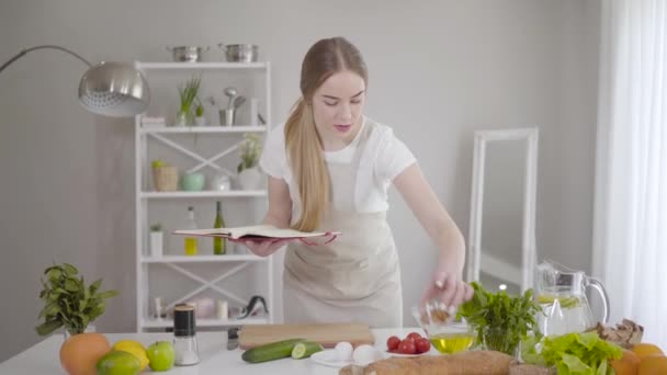Positive young woman reading book of recipes and checking ingredients for dishes. Smiling Caucasian girl cooking dinner at home. Culinary, cuisine, healthy eating, homemade food, lifestyle. — Stock Video