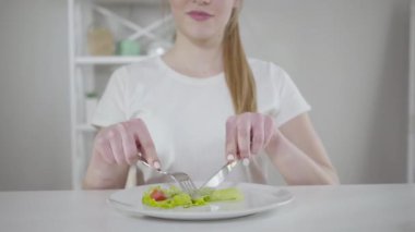 Tanımlanamayan neşeli beyaz kadın sebze salatası yiyor ve gülümsüyor. Genç ve güzel bir kız, salatalığı bıçakla kesip ağzına sokuyor. Sağlıklı yaşam tarzı, diyet, kalori sayımı.