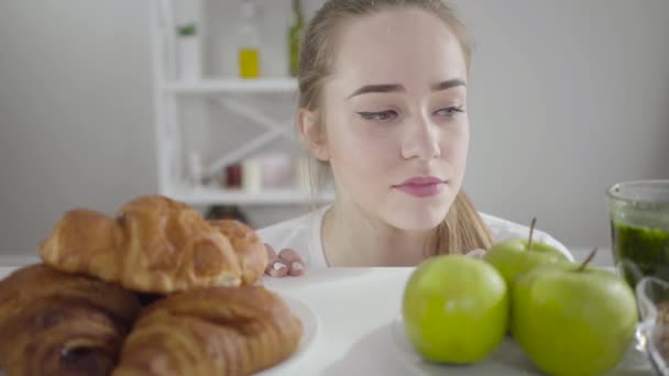 Retrato de bela menina magra escolhendo entre maçãs verdes e croissants crostas. Mulher branca jovem decidindo sobre alimentos saudáveis e não saudáveis. Autocontrole, autodisciplina, estilo de vida . — Vídeo de Stock