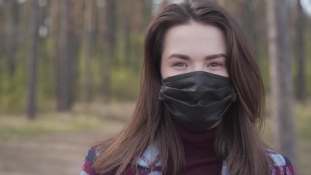 Close-up portrait of beautiful brunette Caucasian girl with brown eyes in black face mask. Pretty woman standing in forest or park and looking at camera. Covid-19 lockdown, coronavirus quarantine. — Stock Video
