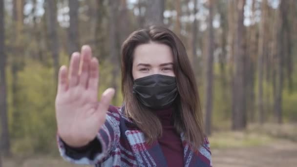 Jonge blanke vrouw met gezichtsmasker met stop gebaar op camera. Portret van een zelfverzekerd brunette meisje met bruine ogen poserend in het bos op coronavirus quarantaine. Covid-19, pandemie, afsluiting.. — Stockvideo