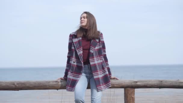 Jeune belle femme caucasienne assise sur une clôture en bois sur la rive de la rivière. Portrait de dame mignonne réfléchie en veste à carreaux et jeans reposant seul près de l'eau par une journée ensoleillée. Mode de vie, loisirs . — Video