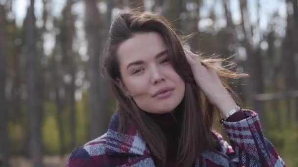 Close-up de atraente jovem posando na floresta de primavera. Retrato de linda morena caucasiana senhora com olhos castanhos fixando o cabelo e sorrindo para a câmera. Natureza, estilo de vida, lazer . — Vídeo de Stock