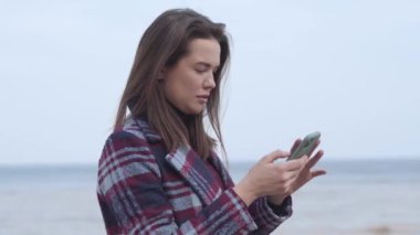 Açık havada akıllı telefon kullanan esmer bir kadının yan görüntüsü. Güneşli bir bahar gününde nehir kenarında dinlenen beyaz bir kızın portresi. Boş zaman, yaşam tarzı, doğa..