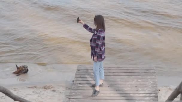 Широкий снимок привлекательной белой девушки, делающей селфи стоя на пирсе. Брюнетка, позирующая на берегу реки или озера. Радость, тысячелетия, образ жизни . — стоковое видео