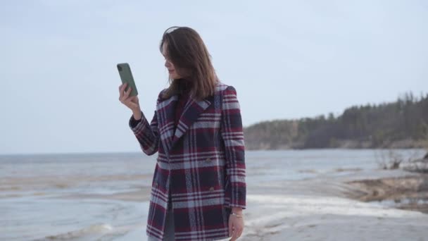 Tiro medio de la morena sonriente chica caucásica grabación de vídeo selfie en el teléfono inteligente al aire libre. Mujer milenaria positiva descansando en la orilla del río arenoso. Ocio, estilo de vida, alegría, naturaleza, blogging . — Vídeos de Stock