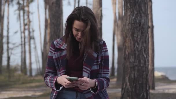 Porträtt av koncentrerad vit flicka med hjälp av smartphone i skogen. Brunett tusenårig kvinna surfa internet utomhus. Trådlös teknik, fritid, livsstil, natur. — Stockvideo