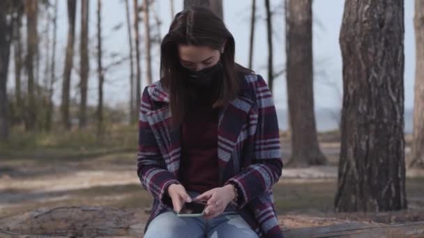 Joven morena mujer caucásica en máscara de cuero sentado en el bosque y el uso de teléfono inteligente. Retrato de niña milenaria navegando por Internet al aire libre. Estilo de vida, comunicación inalámbrica, tecnología moderna . — Vídeo de stock