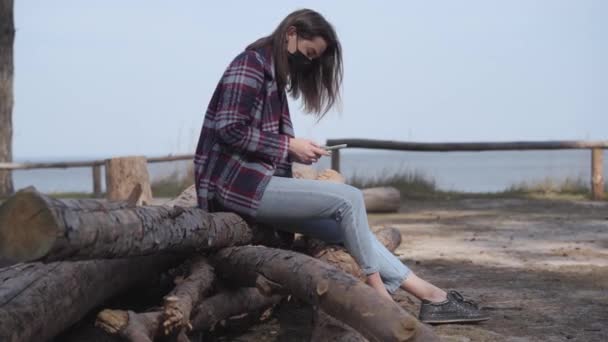 Boční pohled na brunetku v masce pomocí telefonu, jak sedí na kládě vedle řeky. Mladá běloška odpočívá venku na Covid-19. Koronavirová pandemie, jarní volný čas, životní styl. — Stock video