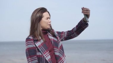 Neşeli beyaz kadın konuşuyor ve dışarıda selfie kamerasına el sallıyor. Güneşli bir günde nehir kıyısında video kaydeden pozitif kadın blogcu portresi. Günlükler, yaşam tarzı, binyıllar, doğa..