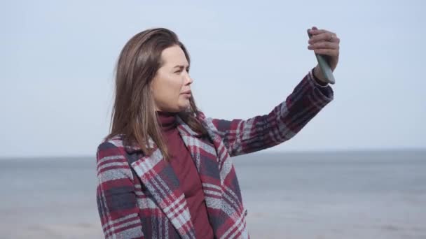 Alegre mujer caucásica hablando y saludando a la cámara selfie al aire libre. Retrato de una bloguera positiva grabando video en la orilla del río en un día soleado. Vlogging, estilo de vida, millennials, naturaleza . — Vídeos de Stock