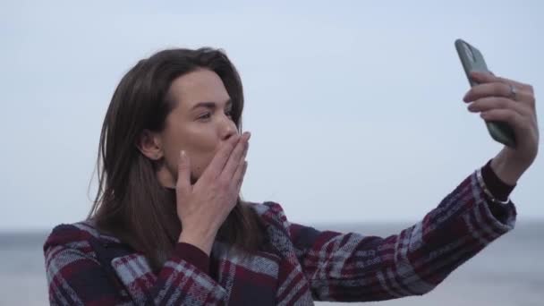 Detailní záběr veselé brunetky, která posílá vzdušný polibek na selfie kameru. Mladá pozitivní běloška fotí na břehu řeky. Radost, životní styl, volný čas, milénia. — Stock video