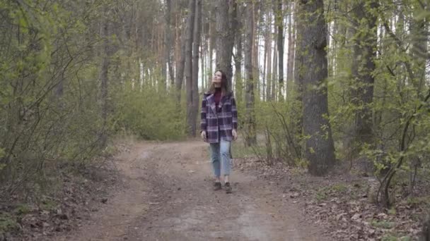 Encantadora mujer caucásica caminando por el callejón del bosque hasta la cámara. Retrato de una hermosa morena vestida con ropa casual paseando al aire libre en el día de primavera u otoño. Ocio, estilo de vida, alegría, relajación — Vídeos de Stock