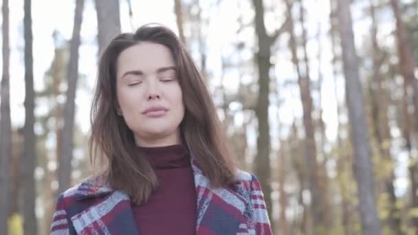 Cámara acercándose a la encantadora cara de la hermosa morena mujer caucásica posando en el bosque. Joven chica de ojos marrones de pie en la luz del sol al aire libre. Naturaleza, ocio, alegría, estilo de vida, belleza . — Vídeo de stock