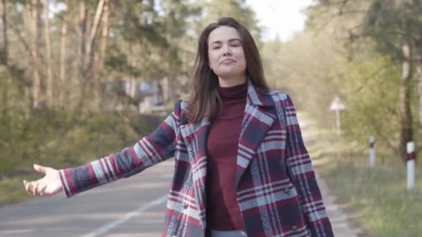Portré aranyos kaukázusi barna lány stoppolás kis úton az erdőben. Pozitív fiatal nő kockás kabátban utazik. Életmód, szabadság, ökoturizmus. — Stock videók