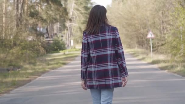 Bakåt bild av självsäker vit brunett kvinna promenader längs tom väg i solig skog. Millennial kvinnlig turist njuter av dagen utomhus. Fritid, lycka, livsstil, fritid. — Stockvideo