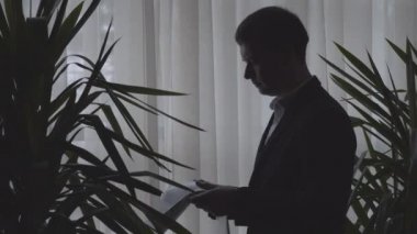 Sabahın erken saatlerinde elinde gazeteyle pencerenin önünde duran yetişkin ciddi iş adamının yan görüntüsü. Takım elbiseli kendine güvenen adam son iş haberlerini okuyor. Yaşam tarzı, aşırı çalışma, başarı, bilgi