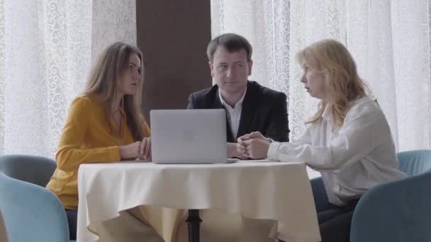 Três empresários caucasianos sérios sentados à mesa com laptop e conversando. Homem profissional e mulheres discutindo a cooperação no café. Parceria, trabalho em equipe, estilo de vida, sucesso . — Vídeo de Stock