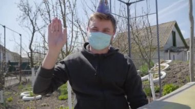 Yüzünde maske ve parti şapkası olan neşeli beyaz adam kameraya el sallıyor. Covid-19 tecridinde doğum gününü kutlayan genç adamın portresi. Karantina, tecrit, yaşam tarzı, eğlence.