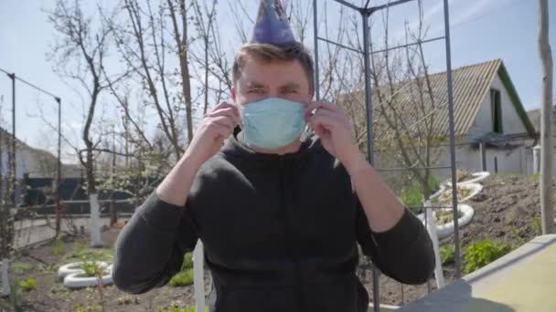 Retrato de un hombre caucásico barbudo serio quitándose la máscara facial y mirando a la cámara. Morena joven en sombrero de fiesta celebrando vacaciones en el chat en línea. Covid-19 cuarentena pandémica, estilo de vida . — Vídeos de Stock