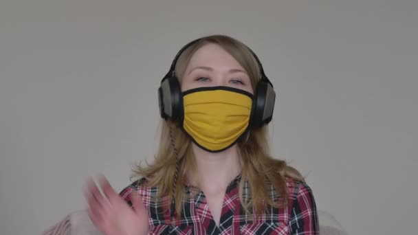 Portrait of joyful young woman in yellow face mask and headphones greeting friends or colleagues in online chat. Blond Caucasian girl waving hello at camera. Remote communication, lockdown, lifestyle. — Stock Video