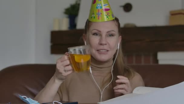 Joyful woman clinking tea cup at camera and talking. Portrait of gorgeous Caucasian lady in party hat on online celebration. Lockdown, quarantine, self-isolation, remote communication, lifestyle. — Stock Video