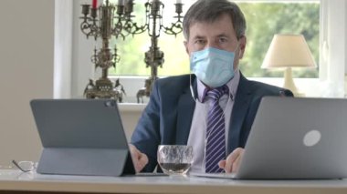 Masada dizüstü bilgisayar ve tabletle oturan, kendine güvenen beyaz maskeli bir işadamı ve biriyle aynı fikirde. Covid-19 karantinasında internet üzerinden iletişim kuran ciddi yetişkin bir adamın portresi.