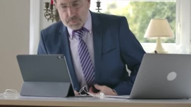 Uyku pantolonlu, ceketli ve kravatlı yetişkin bir beyaz adam masaya geliyor, kulaklık takıyor ve iş arkadaşlarıyla online sohbete başlıyor. Gözlüklü yakışıklı iş adamı tecritte çalışıyor..