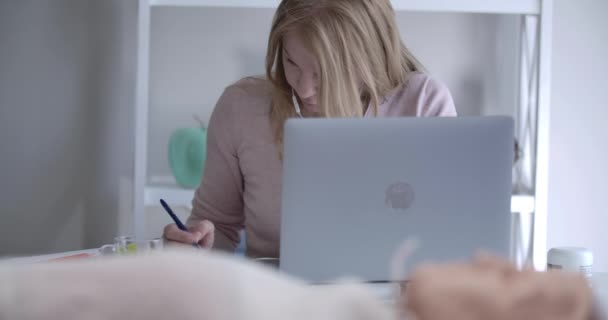 Poważna blondynka, biała kobieta, pisząca notatki przy stole z laptopem jako manekinem do nauki pierwszej pomocy, leżąca na pierwszym planie. Doktor przygotowuje się do egzaminu. Kino 4k ProRes kwatera główna. — Wideo stockowe