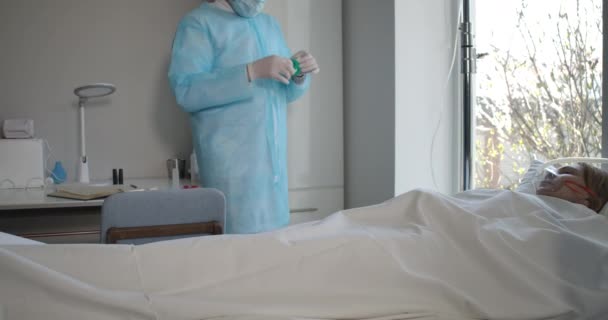 Человек в маске и защитных перчатках заботится о положительном пациенте Ковид-19 в палате больницы. Кавказский врач настраивает капельницу и уходит. Пандемия, здравоохранение, болезни. Штаб-квартира кинотеатра 4k ProRes . — стоковое видео