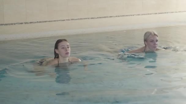 Junge und erwachsene kaukasische Frauen schwimmen im Pool des Hotelkomplexes und unterhalten sich. Glückliche reiche Kundinnen, die sich im luxuriösen Wellnessbereich ausruhen. Freizeit, Wohlstand, Lebensstil, Erfolg. — Stockvideo