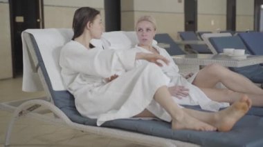 Spa otelindeki güneşli yataklarda dinlenen kendine güvenen genç beyaz kadınlar. Beyaz bornozlu sarışın ve esmer kadınlar konuşuyor. Bayan müşteriler eğlencenin tadını çıkarıyor. Zenginlik, yaşam tarzı, neşe, lüks.