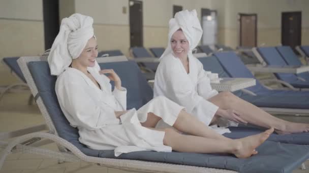 Две веселые богатые дамы в белых халатах и полотенцах для волос смеются и болтают в роскошном спа-курорте. Позитивные молодые кавказские женщины отдыхают в роскошном отеле на отдыхе. Стиль жизни, досуг . — стоковое видео