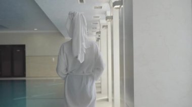 Kafkasyalı bir kadının, spa otelinin koridorunda yürüyüp etrafa bakınışının arka görüntüsü. Beyaz bornoz ve saç havlusuyla lüks bir otelde dinlenen başarılı zengin kadın. Yaşam biçimi.