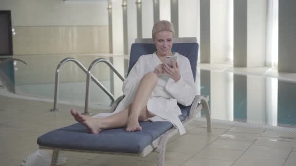 Amplio plano de rubia sonriente mujer caucásica tumbada en la cama de sol en el hotel spa y usando un teléfono inteligente. Señora adulta segura navegando por Internet y descansando en vacaciones. Riqueza, ocio, estilo de vida . — Vídeo de stock