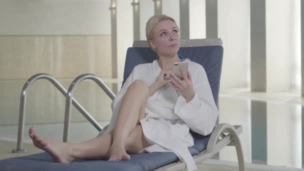 Retrato de mujer rubia caucásica adulta usando teléfono y sonriendo. Encantadora joven descansando en la cama de sol en el hotel spa de vacaciones. Lujo, estilo de vida, riqueza, relajación . — Vídeos de Stock