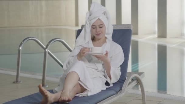 Retrato de la joven encantadora chica caucásica descansando en el spa de vacaciones. Mujer hermosa feliz en albornoz y toalla de pelo tumbado en la tumbona con taza de café y sonriendo. Alegría, ocio, lujo, estilo de vida . — Vídeo de stock