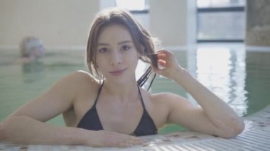 Havuz kenarında, arka planda yüzen tanınmayan sarışın bir kadın gibi poz veren güzel beyaz kız portresi. Lüks spa otelinde kameraya gülümseyen esmer genç bayan. Yaşam tarzı, zenginlik.