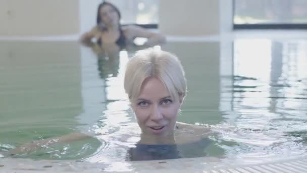Detailní záběr pozitivní blonďaté šedooké ženy pózující u bazénu s uvolněnou brunetkou, která odpočívá v pozadí. Portrét půvabné bělošky, která si užívá dovolenou v luxusním hotelu. — Stock video
