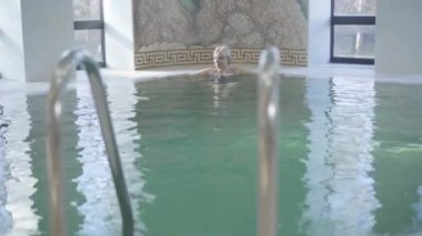 Sarışın, gülümseyen bir kadın yüzme havuzundan geçip gidiyor. Sabahları spa otelinde eğlenen beyaz bir kadının portresi. Boş zaman, turizm, güzellik, neşe.
