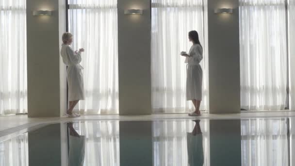 Extreem wijd beeld van twee prachtige blanke vrouwen die 's morgens koffie drinken bij het zwembad. Jonge mooie dames in witte badjassen genieten van een zonnige dag in het kuuroord. Lifestyle, luxe. — Stockvideo