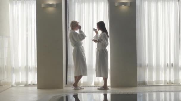 Vista lateral de dos mujeres caucásicas hermosas delgadas que beben café en la piscina en la mañana. Amplio tiro de señoras ricas seguras en batas blancas disfrutando los fines de semana en el balneario. Alegría, ocio . — Vídeo de stock