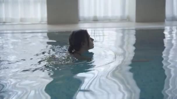 Vista laterale della giovane ragazza caucasica che nuota nel bacino interno. Macchina fotografica a seguito di donna bruna fiduciosa in piscina, passando attrezzature in primo piano. Tempo libero, stile di vita, sport, fiducia . — Video Stock